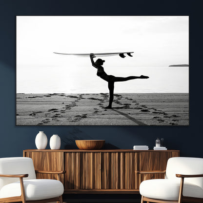 A woman balances a surfboard on her head while striking a yoga pose on the beach, silhouetted against a calm ocean horizon.
