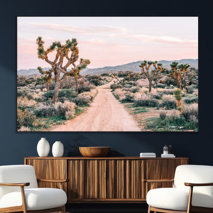A dusty road winding through Joshua Tree National Park, framed by iconic twisted trees and warm desert tones under a soft pastel sky.