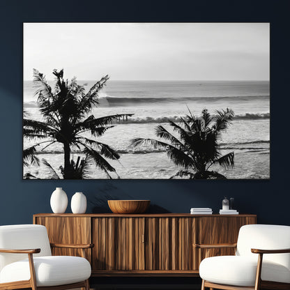 Black and white view of ocean waves crashing on a tropical coastline, framed by silhouetted palm trees under a vast, open sky.