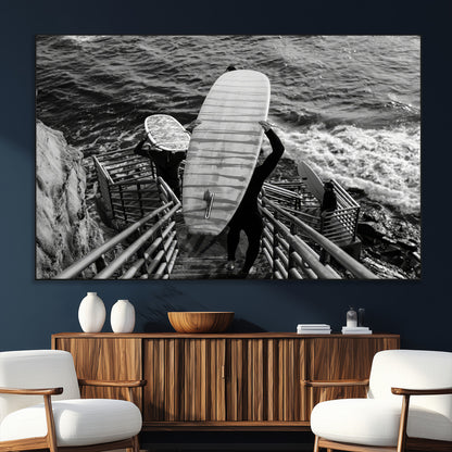 Black and white photo of surfers carrying boards down a rugged cliffside stairway, capturing a bold and adventurous coastal scene.