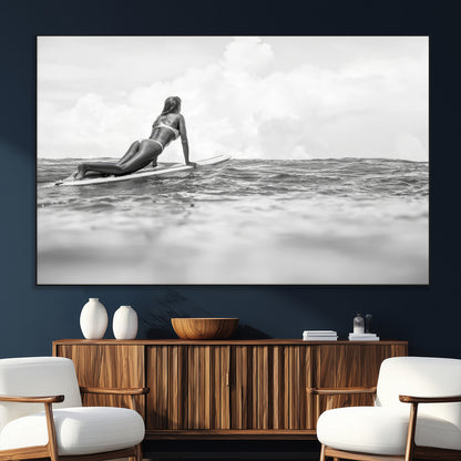 A black and white ocean scene featuring a surfer girl waiting on her board, gazing at the horizon, capturing a tranquil and adventurous coastal moment.