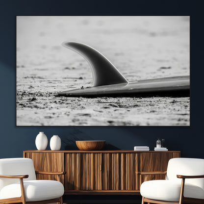 A black and white close-up of a surfboard fin resting on the sandy shore, highlighting minimalist beach aesthetics and classic surf culture.