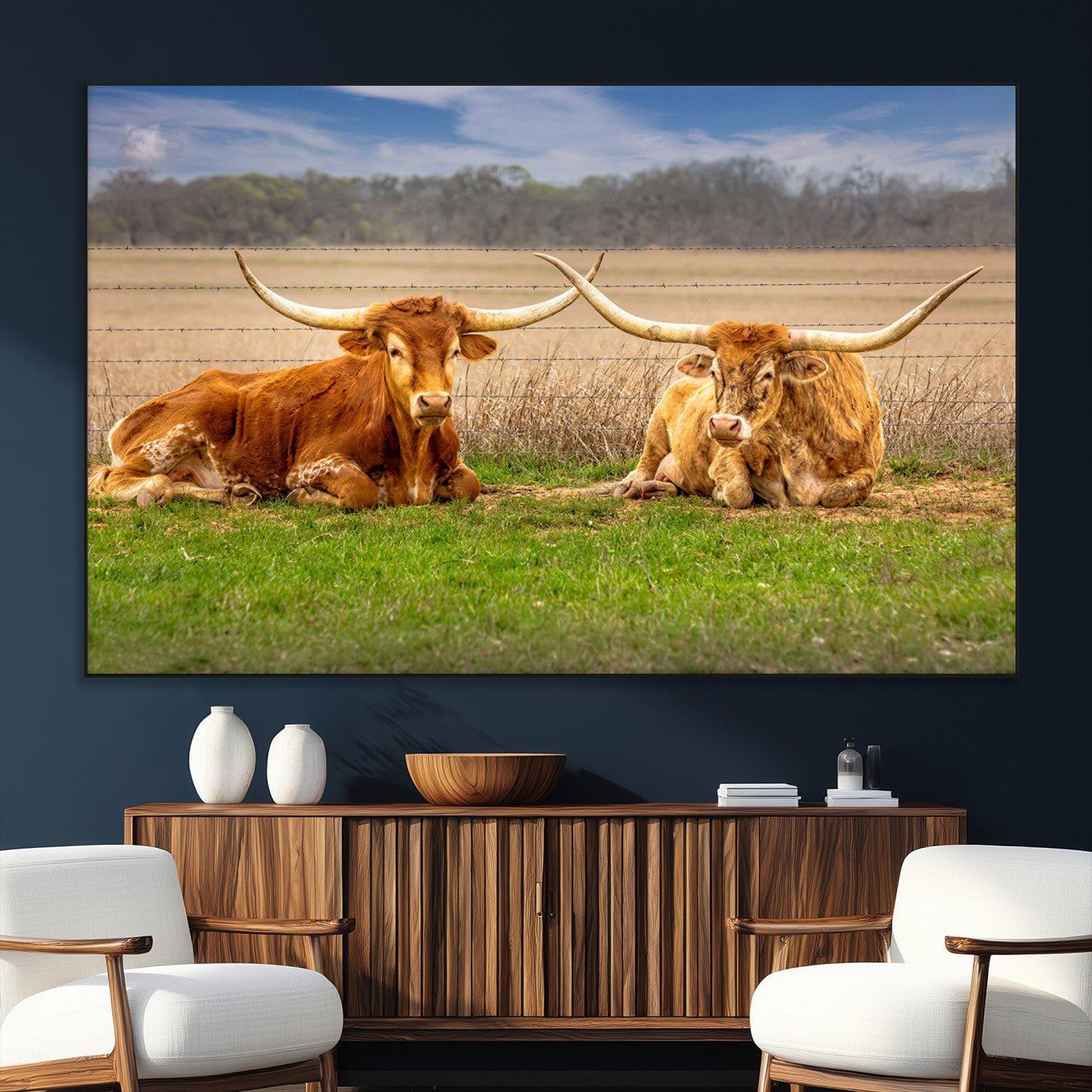 Two longhorn cows lying on green grass in front of a golden field and trees under a blue sky, perfect for rustic country wall decor.