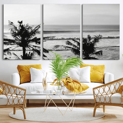 Black and white view of ocean waves crashing on a tropical coastline, framed by silhouetted palm trees under a vast, open sky.