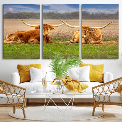 Two longhorn cows lying on green grass in front of a golden field and trees under a blue sky, perfect for rustic country wall decor.