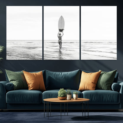 Black and white image of a female surfer holding a surfboard above her head while walking toward the ocean, creating a minimalist and bold coastal aesthetic.