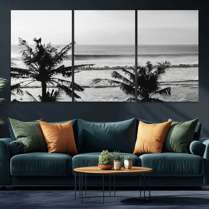 Black and white view of ocean waves crashing on a tropical coastline, framed by silhouetted palm trees under a vast, open sky.