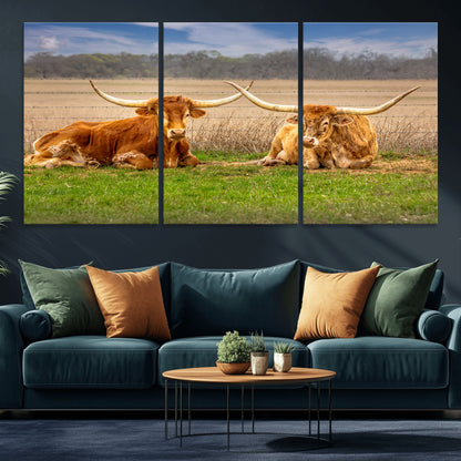 Two longhorn cows lying on green grass in front of a golden field and trees under a blue sky, perfect for rustic country wall decor.