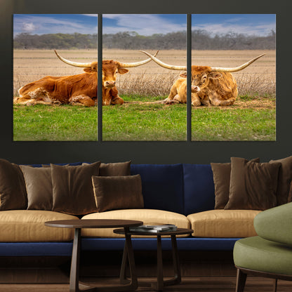 Two longhorn cows lying on green grass in front of a golden field and trees under a blue sky, perfect for rustic country wall decor.