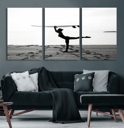 A woman balances a surfboard on her head while striking a yoga pose on the beach, silhouetted against a calm ocean horizon.