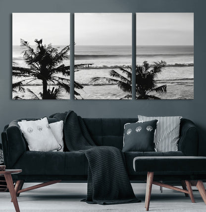 Black and white view of ocean waves crashing on a tropical coastline, framed by silhouetted palm trees under a vast, open sky.