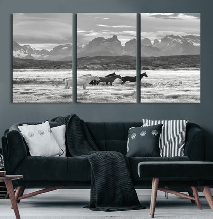A black and white landscape of wild horses grazing in a vast grassland with towering mountains in the background, creating a moody and rustic western aesthetic.