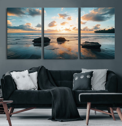 A serene beach at sunset with vibrant clouds, calm water, and rocks in the foreground reflecting the warm colors of the setting sun.