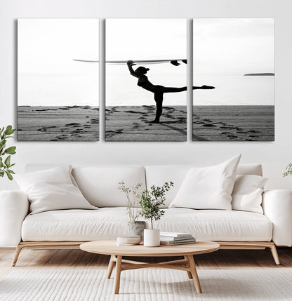 A woman balances a surfboard on her head while striking a yoga pose on the beach, silhouetted against a calm ocean horizon.