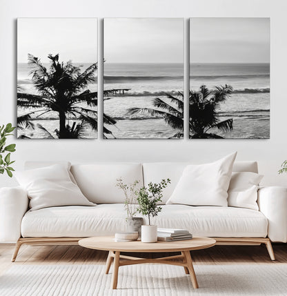 Black and white view of ocean waves crashing on a tropical coastline, framed by silhouetted palm trees under a vast, open sky.