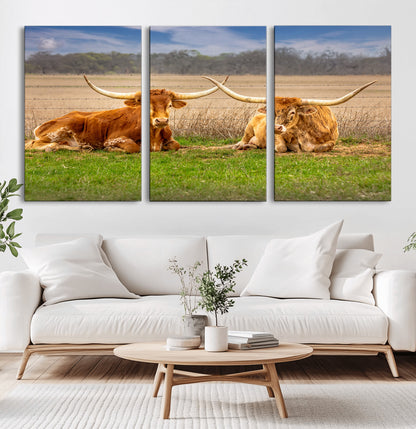 Two longhorn cows lying on green grass in front of a golden field and trees under a blue sky, perfect for rustic country wall decor.