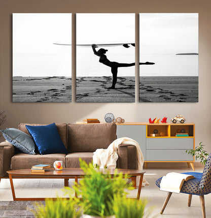 A woman balances a surfboard on her head while striking a yoga pose on the beach, silhouetted against a calm ocean horizon.