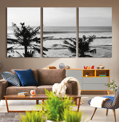 Black and white view of ocean waves crashing on a tropical coastline, framed by silhouetted palm trees under a vast, open sky.