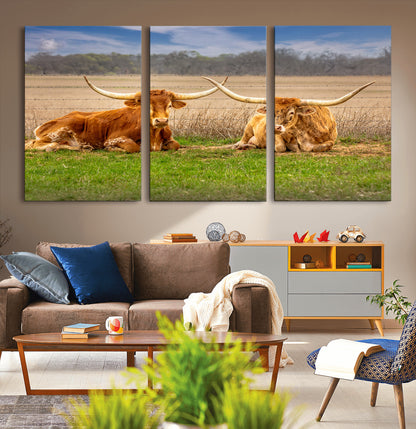 Two longhorn cows lying on green grass in front of a golden field and trees under a blue sky, perfect for rustic country wall decor.