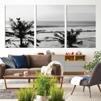 Black and white view of ocean waves crashing on a tropical coastline, framed by silhouetted palm trees under a vast, open sky.